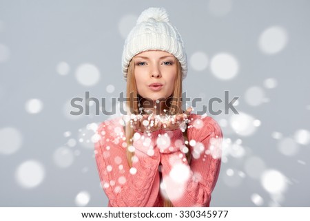 Christmas Girl. Beautiful caucasian woman wearing winter clothing hat blowing snow at you, looking at camera, over gray background