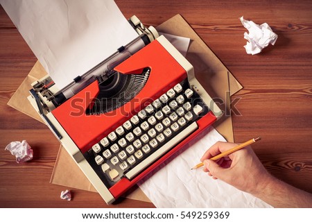 Top view of red vintage typewriter with white blank paper sheet with male hand holding pencil making drafts, on wooden table