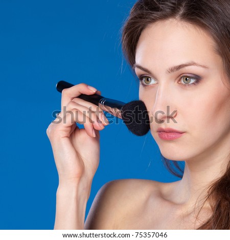 Young pretty female applying mineral powder on her face with the brush, isolated on blue