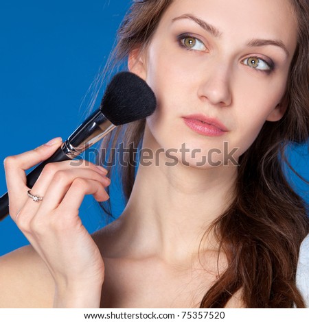 Young pretty female applying mineral powder on her face with the brush, isolated on blue