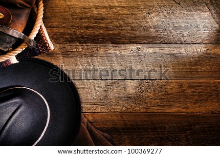 American West rodeo cowboy black felt hat with tools of the trade and authentic brown leather western boots with spur on old weathered ranch barn wood floor as a grunge background