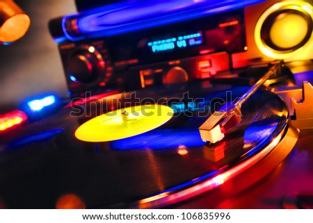 Vinyl record playing hot techno music on an audio DJ turntable with disc jockey sound system equipment in a hip disco dance club lit in colorful party lights effects