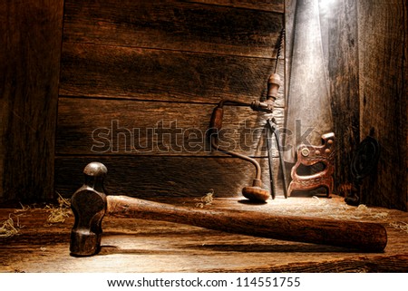 Old antique wood handle blacksmith hammer and used antique carpenter tools with ancient manual drill and crosscut saw in vintage wood working carpentry workshop lit by soft diffused light through dust