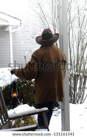 American West legend cowboy with western hat and leather jacket on a cold ranch front porch holding a lit kerosene lantern while contemplating new falling snow in winter weather