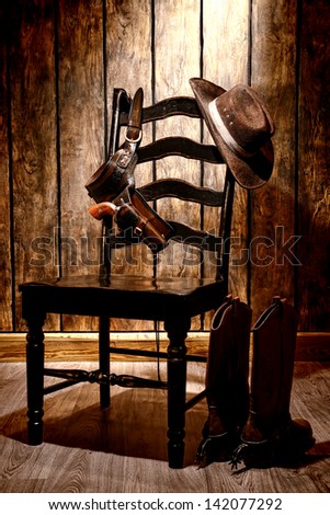 American West legend traditional brown cowboy hat and revolver gun in leather holster hanging on an old black wooden chair with authentic boots on wood floor in a vintage ranch western home