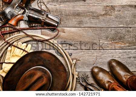 American West legend western cowboy ranching gear still life with old revolver gun in leather holster along lariat lasso and antique hat near boots and spurs on wood board ranch barn floor background