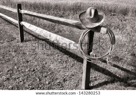 American West rodeo old cowboy hat and wrangling lasso lariat hanging on an old wood fence post on a farm field at a western ranch