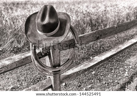 American West rodeo traditional cowboy hat and roping lasso lariat hanging on an old wood fence post near a western ranch field in black and white