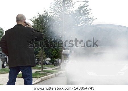 Stranded motorist driver on side of road calling on cell phone for emergency assistance help near broken down car with smoke or steam from hose leak smoking out of open automobile engine compartment