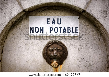Non drinkable water enamel warning sign in French over antique lion head spout on an old public square fountain in France