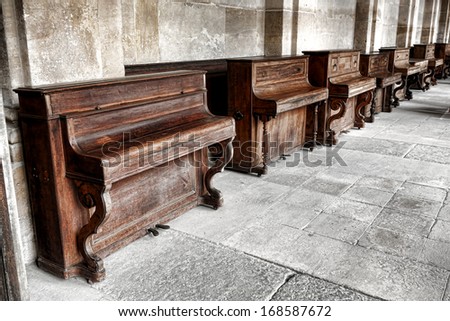 Row of vintage upright study pianos with weathered wood cabinet in an old music school lesson hall with antique stone walls and floors