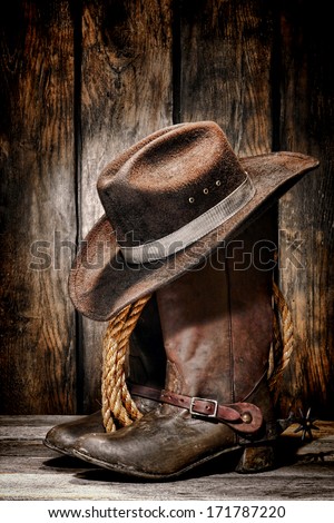 American West Rodeo Cowboy Dirty And Used Black Felt Hat Atop Worn And Old Leather Working Rancher Boots With Vintage Spurs And Ranching Rope In An Antique Ranch Barn