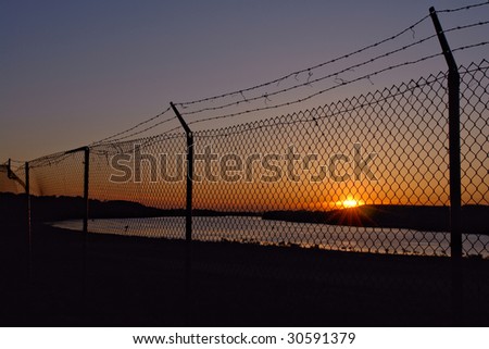 Barb wire on top of chain link fence at the border near a river at sunset