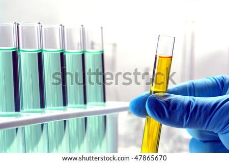 Scientist hand holding a laboratory glass test tubes filled with yellow liquid next to a rack of tubes filled with green chemical solution for an experiment in a science research lab
