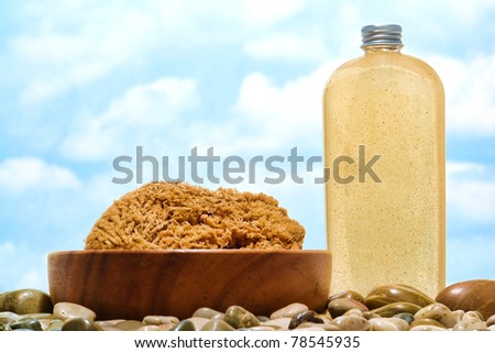 Natural organic bath sponge in a wood bowl and body scrub cleaning wash liquid soap cosmetics bottle over rocks and pebbles on a beach with blue sky
