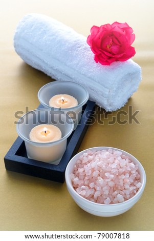 Aromatic bath salts in a ceramic bowl with burning candles in glass holders and rose flower on soft white towel over silky gold surface for pampering relaxation session in a sophisticated modern spa