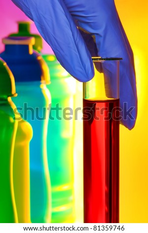 Scientist hand holding a laboratory glass test tube filled with red liquid solution next to colorful bottles for an experiment in a science research lab