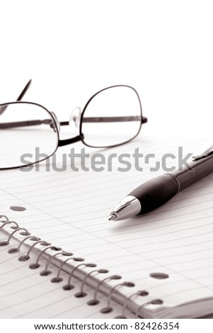 Ballpoint ink writing pen ready for taking notes on a spiral bound study and composition notebook open to a blank ruled paper sheet with reading glasses over white