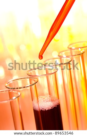 Laboratory pipette with drop of red liquid over glass test tubes causing a chemical reaction inside a tube filled with purple solution for an experiment in a science research lab