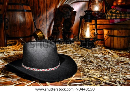 American West rodeo cowboy traditional black felt hat on straw covered wood floor in vintage ranch barn with antique ranching supplies and rancher tools lit by old nostalgic kerosene lantern oil lamps