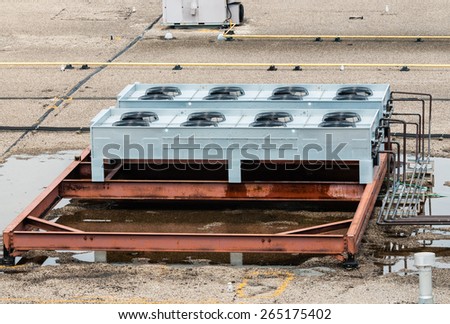 Ventilation fans, pipes, red steel bars, and other industrial machinery on flat roof top. Puddles of standing water are under machinery, and part of girder is wet.