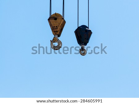Two dirty industrial crane hoist hooks and pulleys hanging by steel cables on pale blue sky.