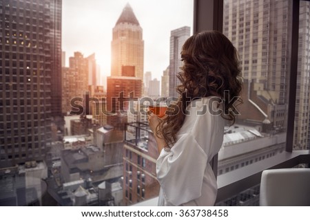 Beautiful woman holding coffee cup and looking to the window in luxury Manhattan penthouse apartments. Good morning after wake up.