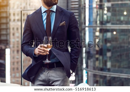 Concept photo of rich people luxury life. Adult successful elegant businessman wearing suit and drinking wine on the rooftop in luxury penthouse in New York City.