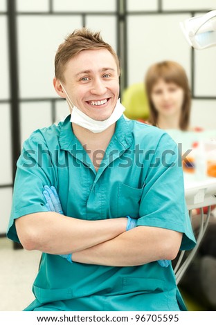 Closeup portrait of young happy doctor in his surgery looking at the viewer, in the background his a female patient