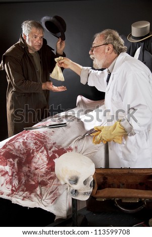 Evil doctor reaches over bloody corpse and pays grave robber, who tips hat. Stage effect with dark background, spot lighting.
