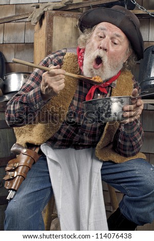 Classic Old West style cowboy with felt hat, grey whiskers, red bandana. He sits on a stool eating beans from a saucepan. Camp cookware and wood shingles in background.
