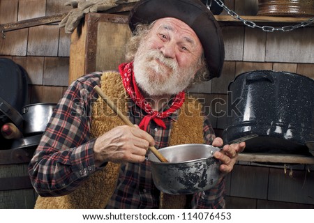 Classic Old West style cowboy with felt hat, grey whiskers, red bandana. He stirs a saucepan with a wooden spoon. Camp cookware and wood shingles in background.