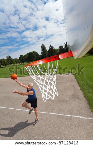 Active retired man shoots hoops outdoors. His dynamic shadow is cast on the court. Background of grass, trees, blue sky, fluffy clouds.  Wide angle looking down from the net. Copy space provided.
