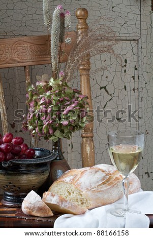 French country style still life with crusty loaf of bread, white wine, flowers and red grapes. Background is rustic chair and painted door. Vertical format and copy space.