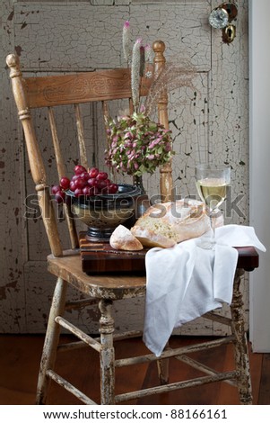 French country style still life with crusty loaf of bread, white wine, flowers and red grapes. Background is rustic chair and painted door. Vertical format and copy space.