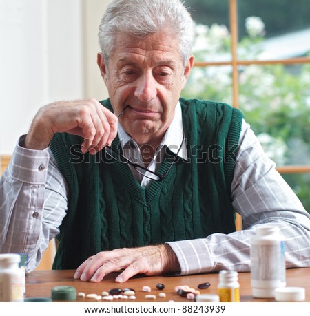 Senior man with glasses in hand looks thoughtfully at many pills on table in front of him. Focus on man. Frontal view, square format, green and white color palette.