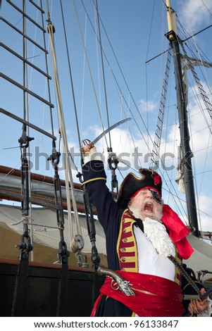 Pirate captain in colorful traditional costume stands on board ship, shouts, and waves his sword. Sailing ship rigging and blue sky in background, vertical layout.