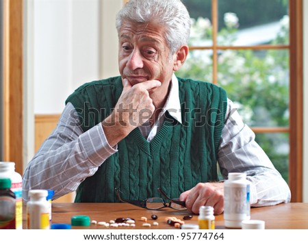 Senior man with glasses on table strokes chin and looks thoughtfully at many pills on table in front of him. Focus on man. Frontal view, green and white color palette.