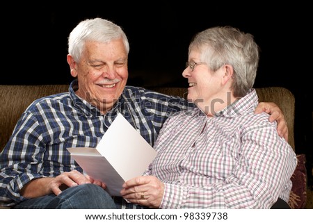 Happy senior married couple enjoy a greeting card, sitting together on a sofa. Black background, horizontal with copy space.