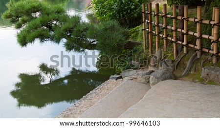 Stone steps lead down to water\'s edge, with pine bough overhanging and reflected in the calm water\'s surface. Traditional Japanese bamboo fence forms grid background. Horizontal landscape.