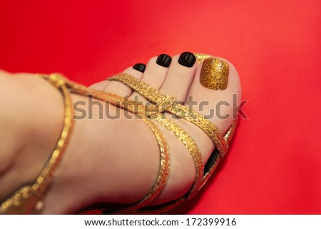 Beautiful luxury pedicure with black and gold brilliant varnish on a red background in gold sandals.
