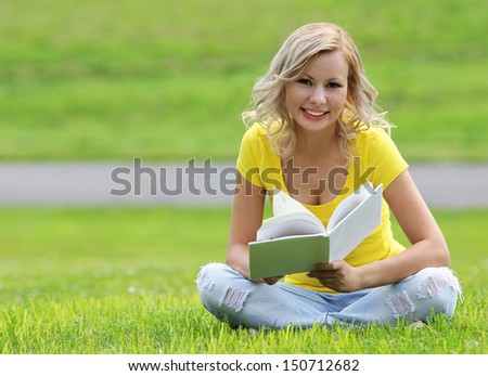 Girl reading the book. Happy blonde beautiful young woman with book sitting on the grass. Outdoor. Looking at the camera