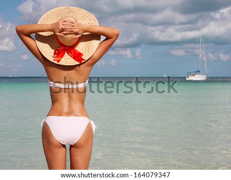 Sexy girl on tropical beach. Beautiful young woman with sun hat and bikini from behind on shore enjoying blue sea. Vacation.