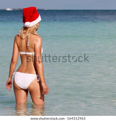 Santa Girl on Tropical Beach. Beautiful blonde young woman in red Christmas Hat and bikini from behind enjoying blue sea. Christmas Vacation. Concept