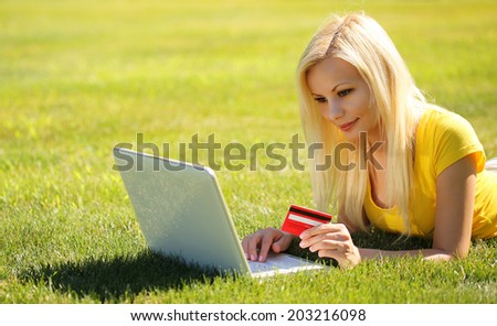 Online Shopping. Smiling Blonde Girl with Laptop using Credit Card and Lying Green Grass.