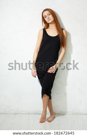 Skinny girl with long legs in black dress standing on a white floor against a white textured wall.