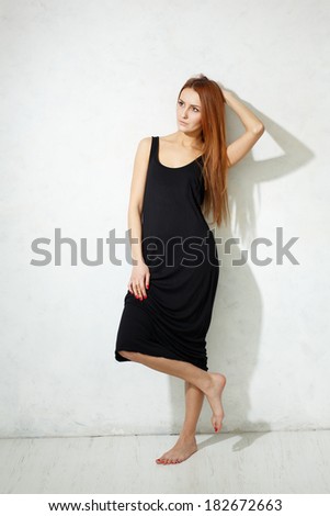 Skinny girl with long legs in black dress standing on a white floor against a white textured wall.
