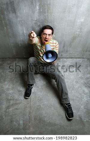 man sits on the floor and screaming into a megaphone, pointing, on the background of gray textured wall.