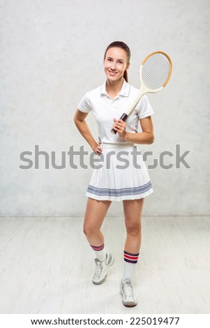 Beautiful girl in tennis dress, standing with a tennis racket in the hands of a white textured wall.
