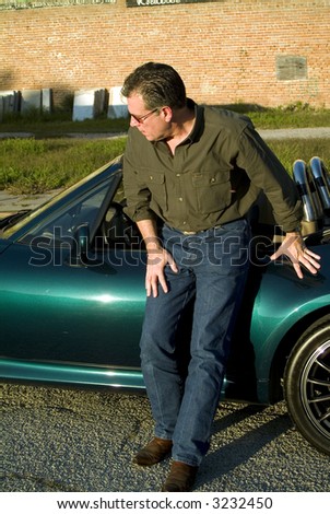 Man inclined slightly looking towards the front of his car as if concerned that his tire may be flat.